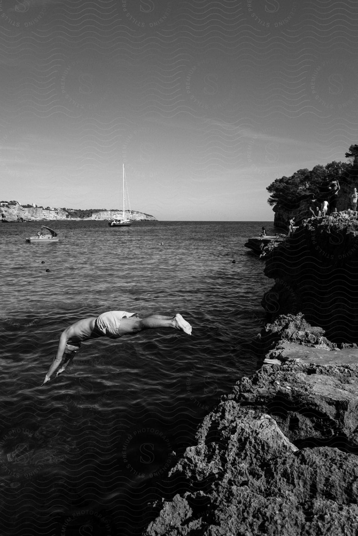 Man diving off rocky edge into mediterranean sea with sailboats in the background