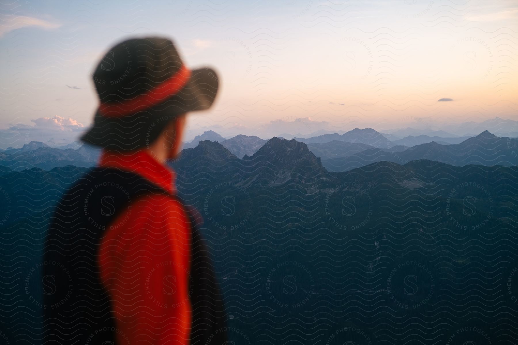 Man wearing a black hat red shirt and black vest looks at distant mountain range during sunset in the alps