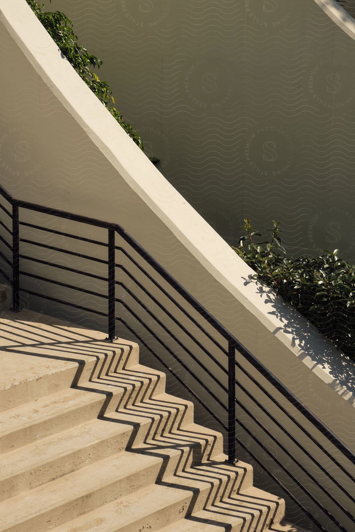 Outdoor staircase railing casts a patterned shadow onto a buildings stairs with a bush behind a wall