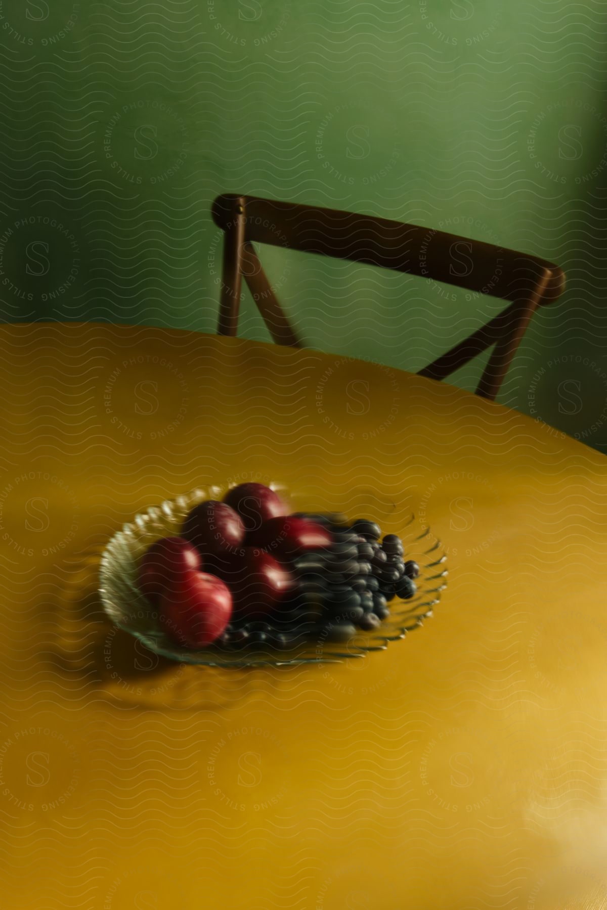 A glass plate of grapes and apples on table with a chair pushed up against the table in a studio with a green wall