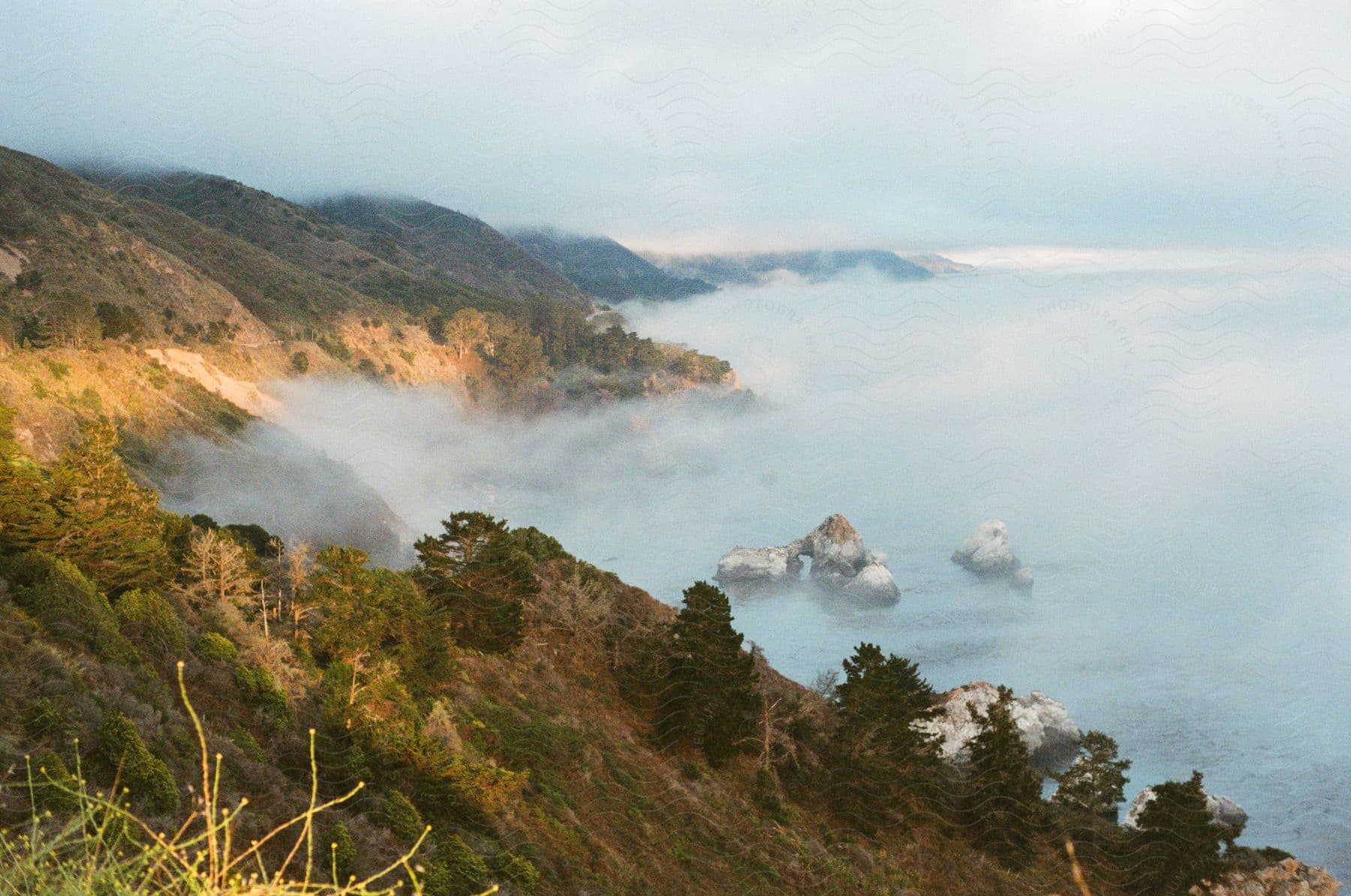 Rolling hills meeting the ocean covered in fog under a cloudy sky