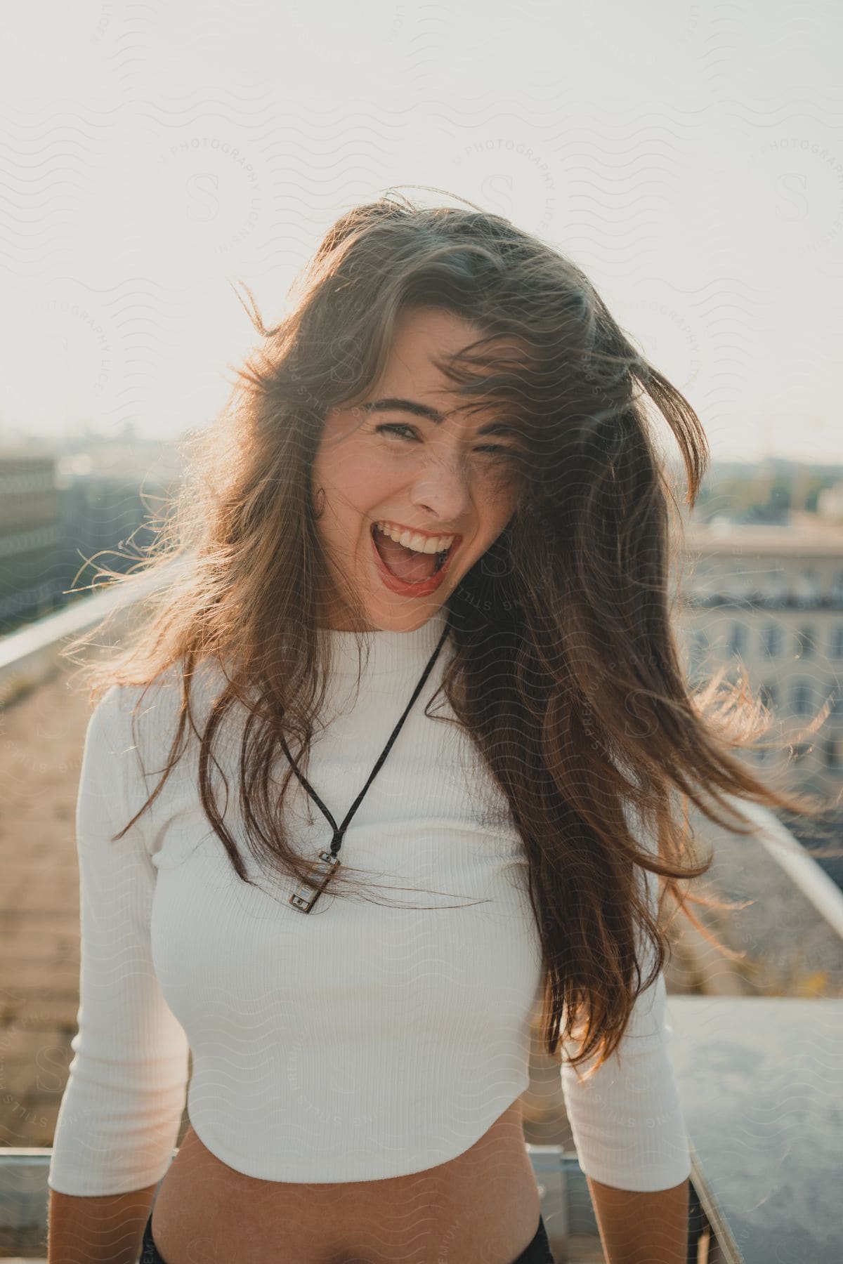 Woman with long hair laughing on a sunny rooftop, wearing a white top and a necklace.