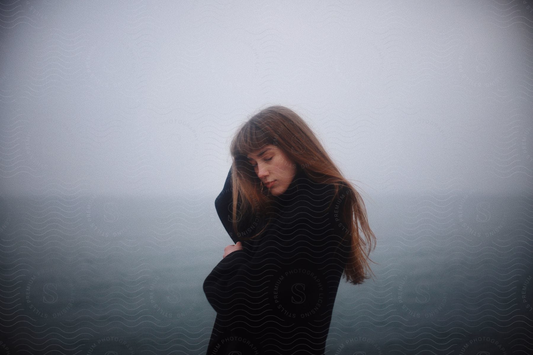Woman with closed eyes stands on cliff against ocean on cloudy day