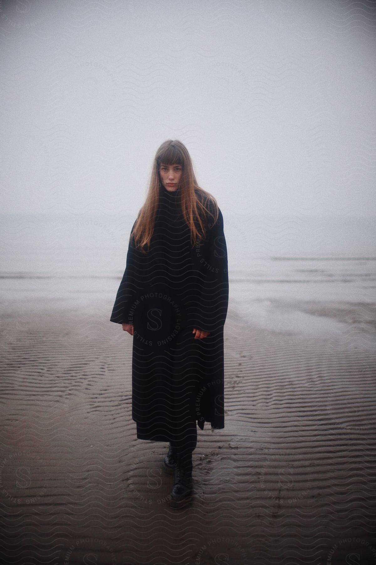 A woman in a long black coat and boots walks along a beach