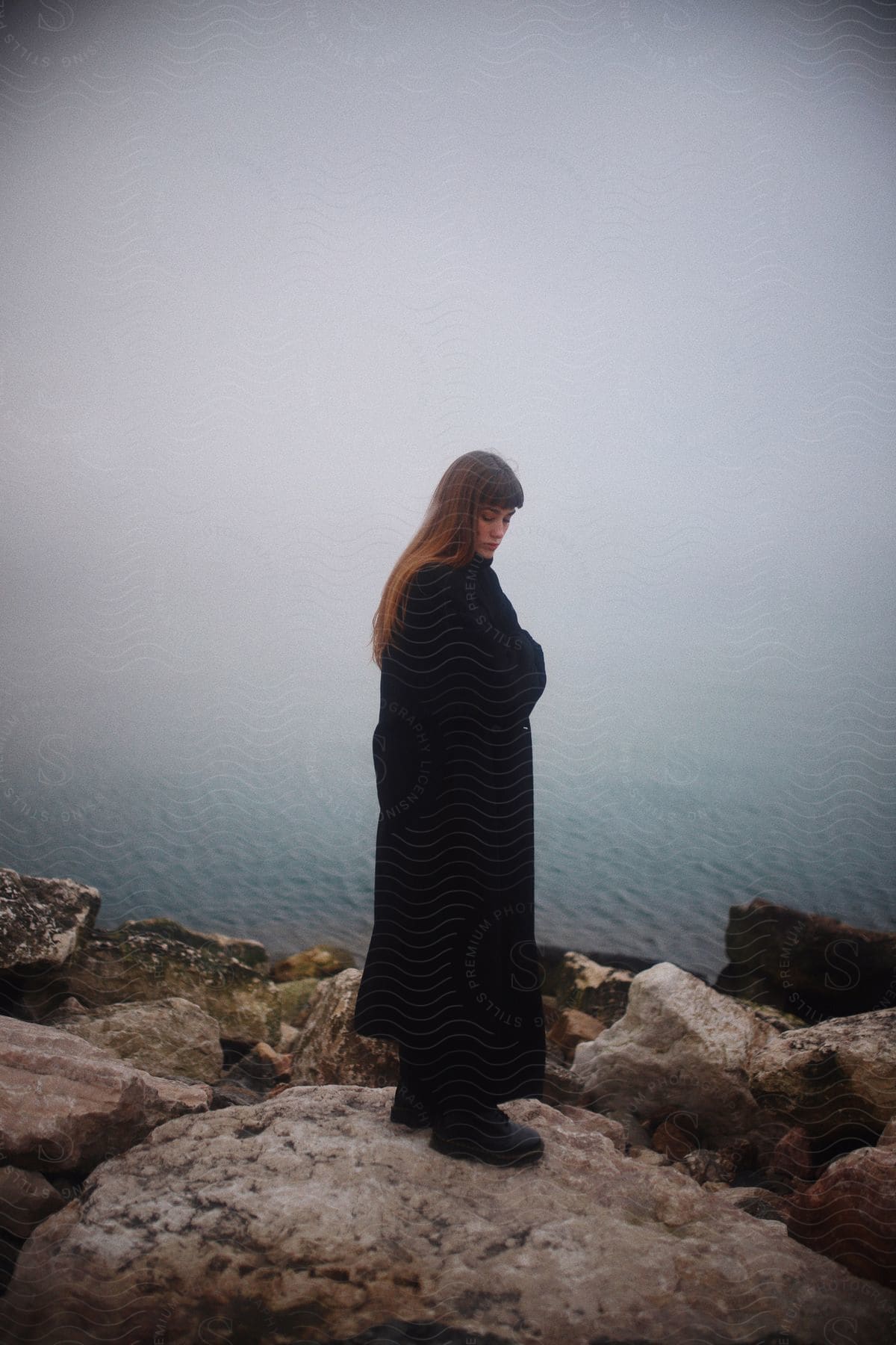 A woman stands on a cliff in a long black coat as the sun sets over a foggy lake