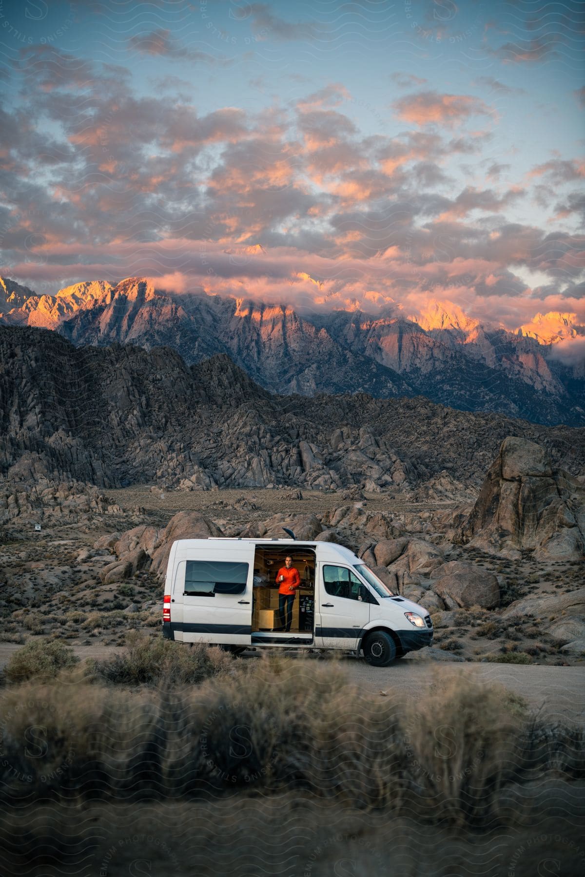 A white van is parked on a desert road with mountains in the distance with the door open and a person standing inside looking out