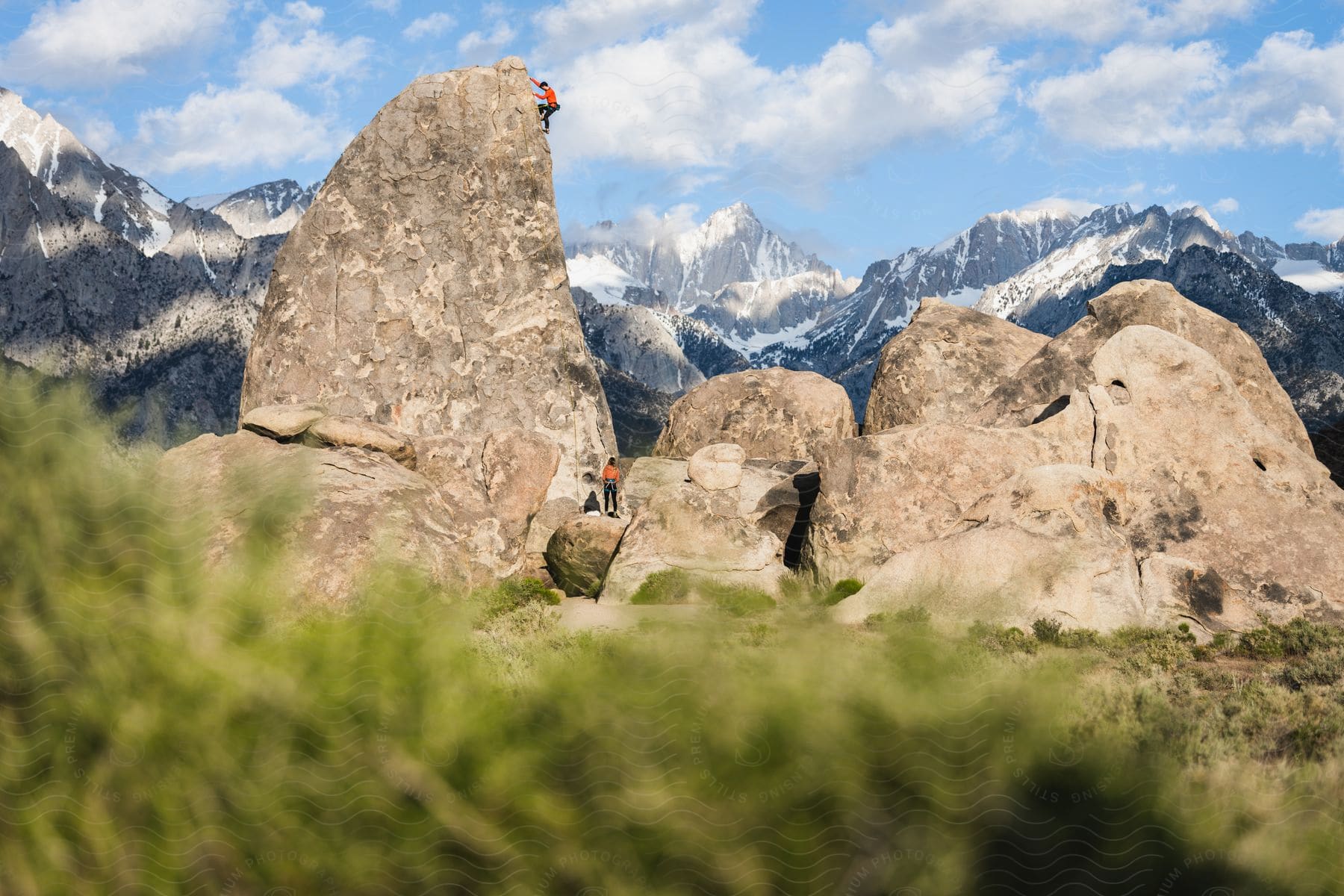 A rock climber ascends a large rock as someone watches from below with mountains in the distance