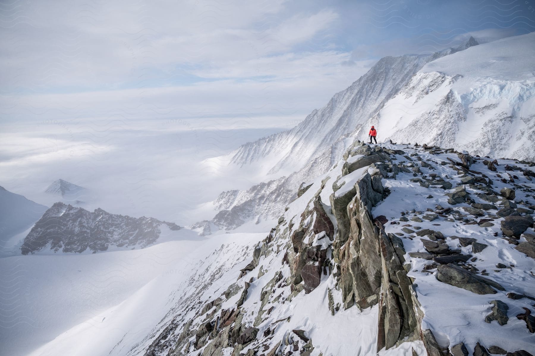 A man stands on a snowcovered mountain with rocky terrain surrounded by distant mountains under a cloudy sky