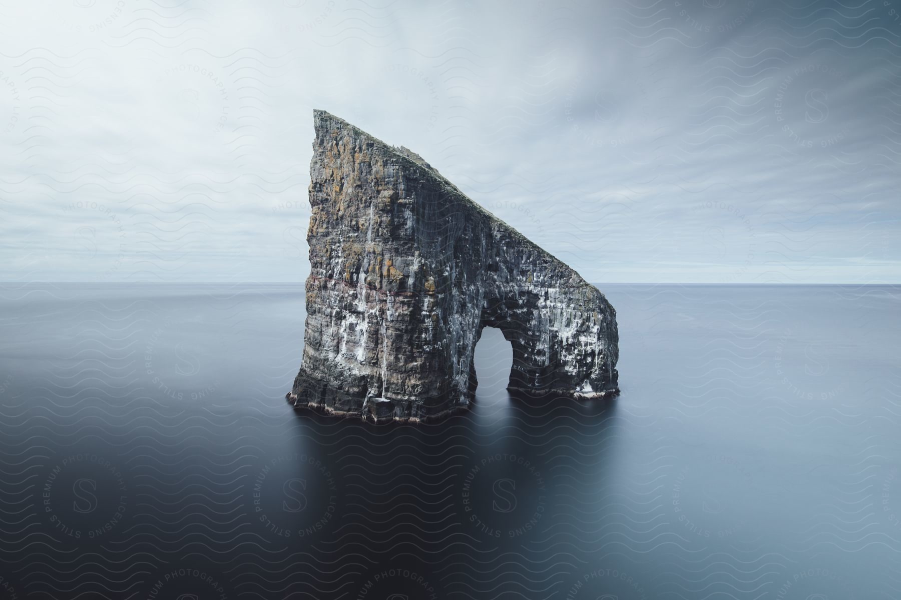 An arch rock formation stands in the water under a cloudy sky