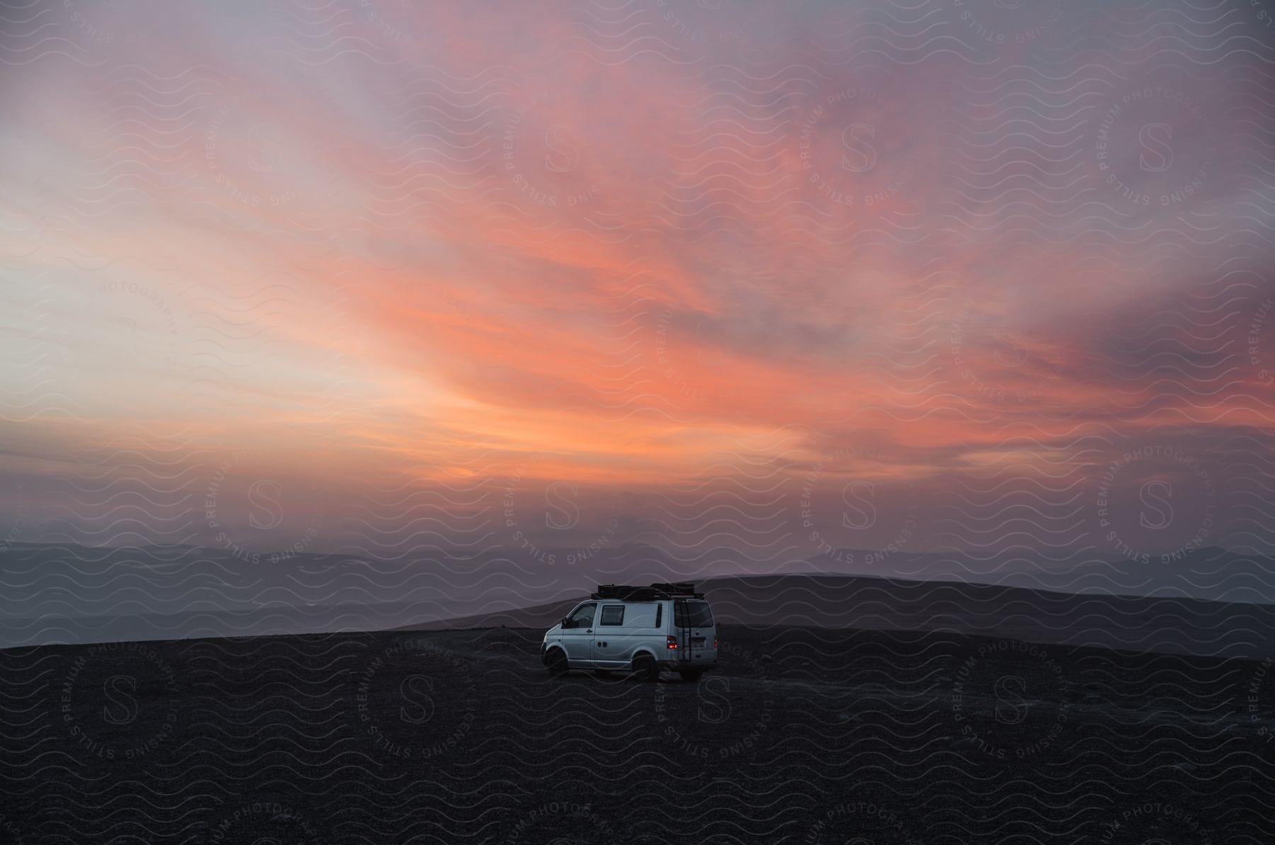 A van drives across a desert with mountains in the distance under a colorful hazy sky at sunset