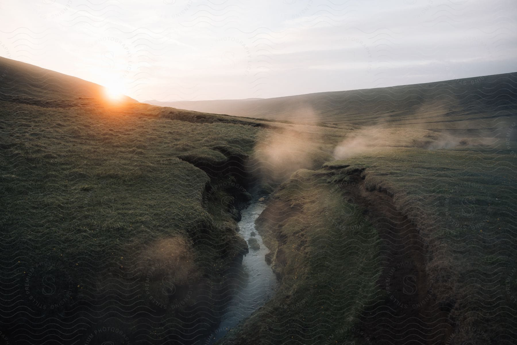 Fog rises over a stream on a hillside as the sun shines brightly over the mountain