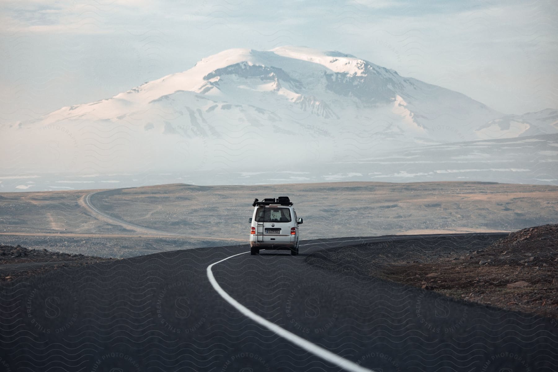 A white van travels on a road toward a snow covered mountain under a cloudy sky