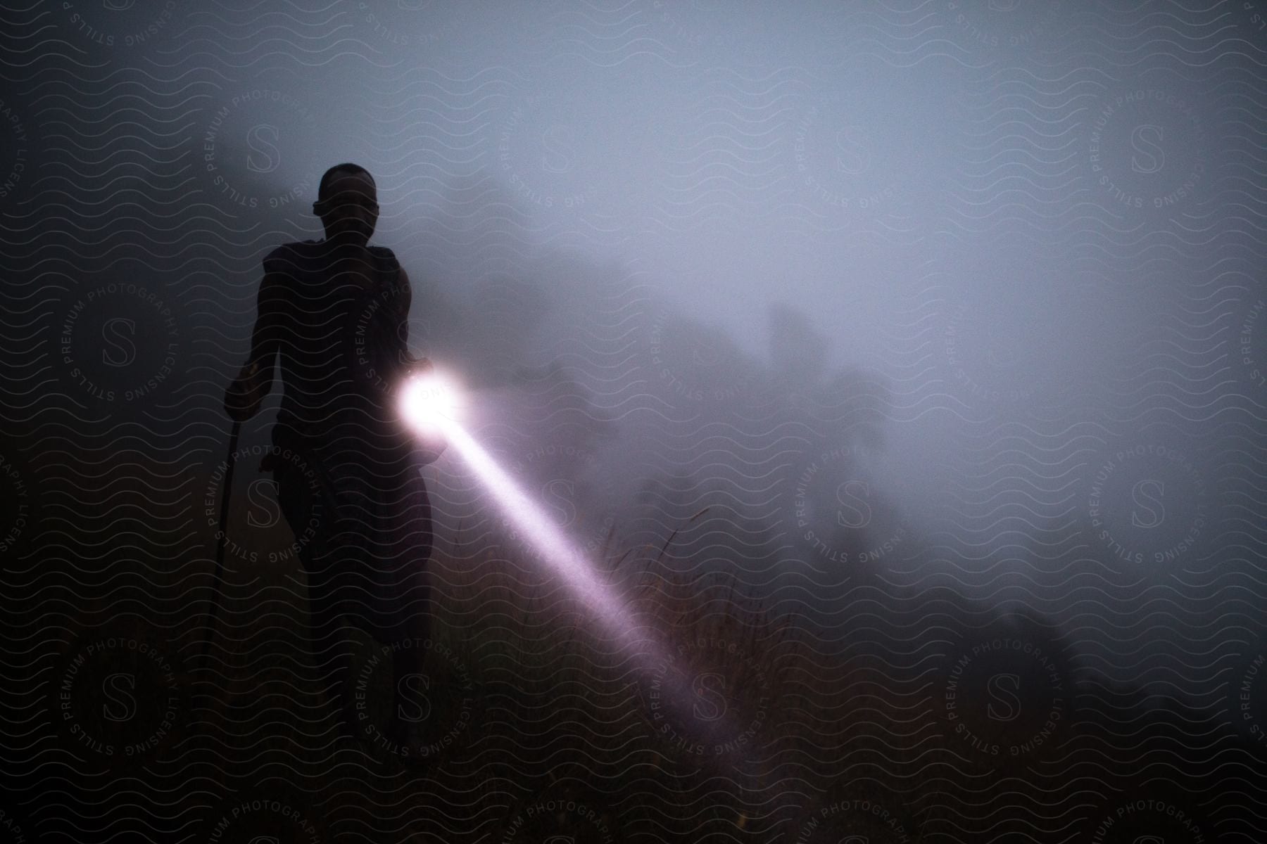 Silhouette of a person walking on a mountainside in thick fog at night holding a flashlight