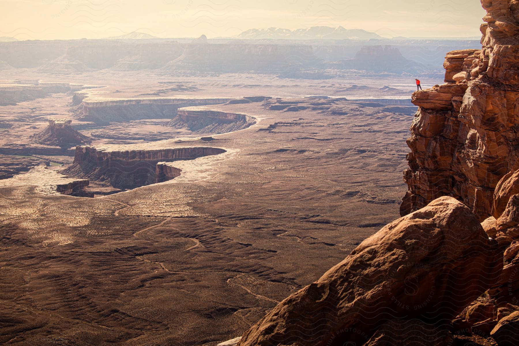 A hiker gazes over a cliff at a panoramic view of canyons mesas and mountains on a hazy day