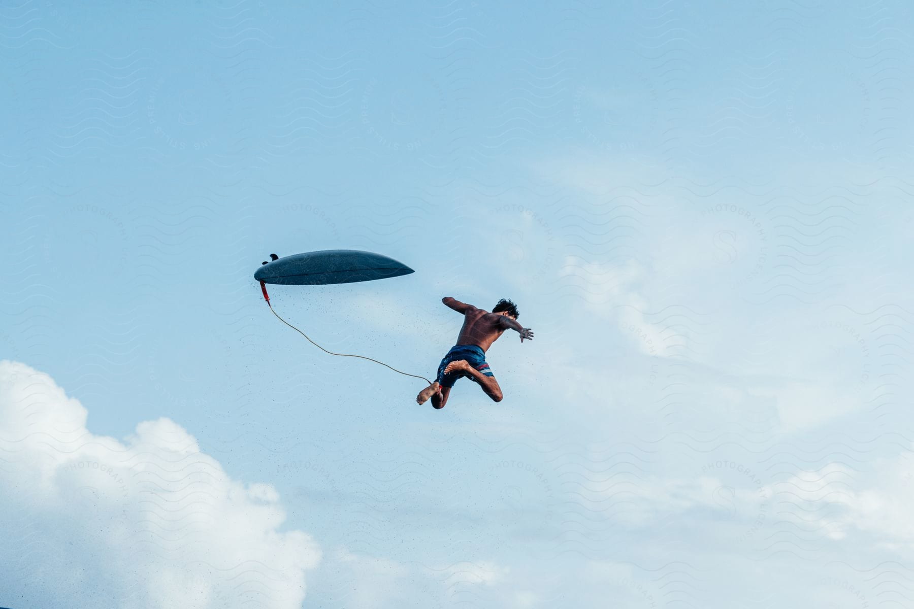 A determined male surfer falls through the air with a surfboard attached to him by an ankle cord against a sky with clouds