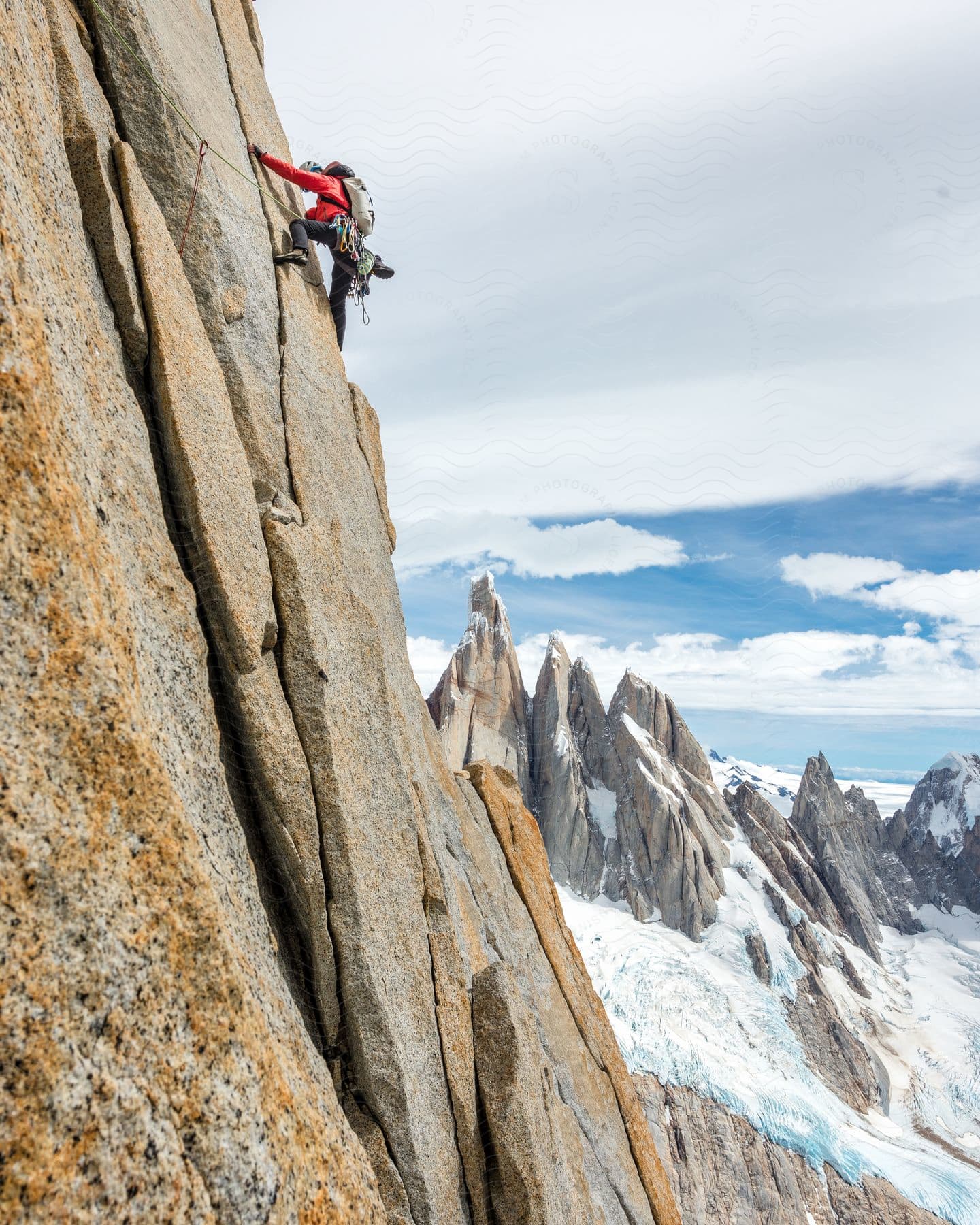 A redjacketed rock climber ascends a cloudy mountainside equipped with helmet rope and guide with snowcapped peaks in the distance