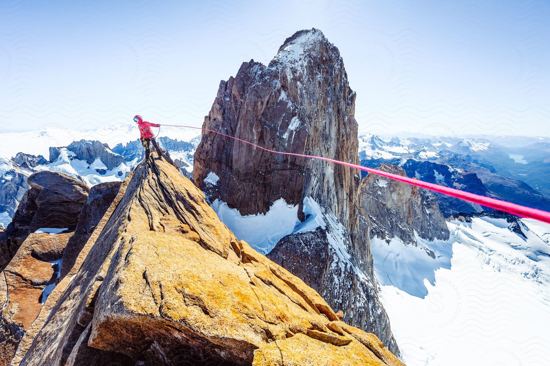 A man stands on a snowcapped mountain with a red guide rope with another mountain in the background