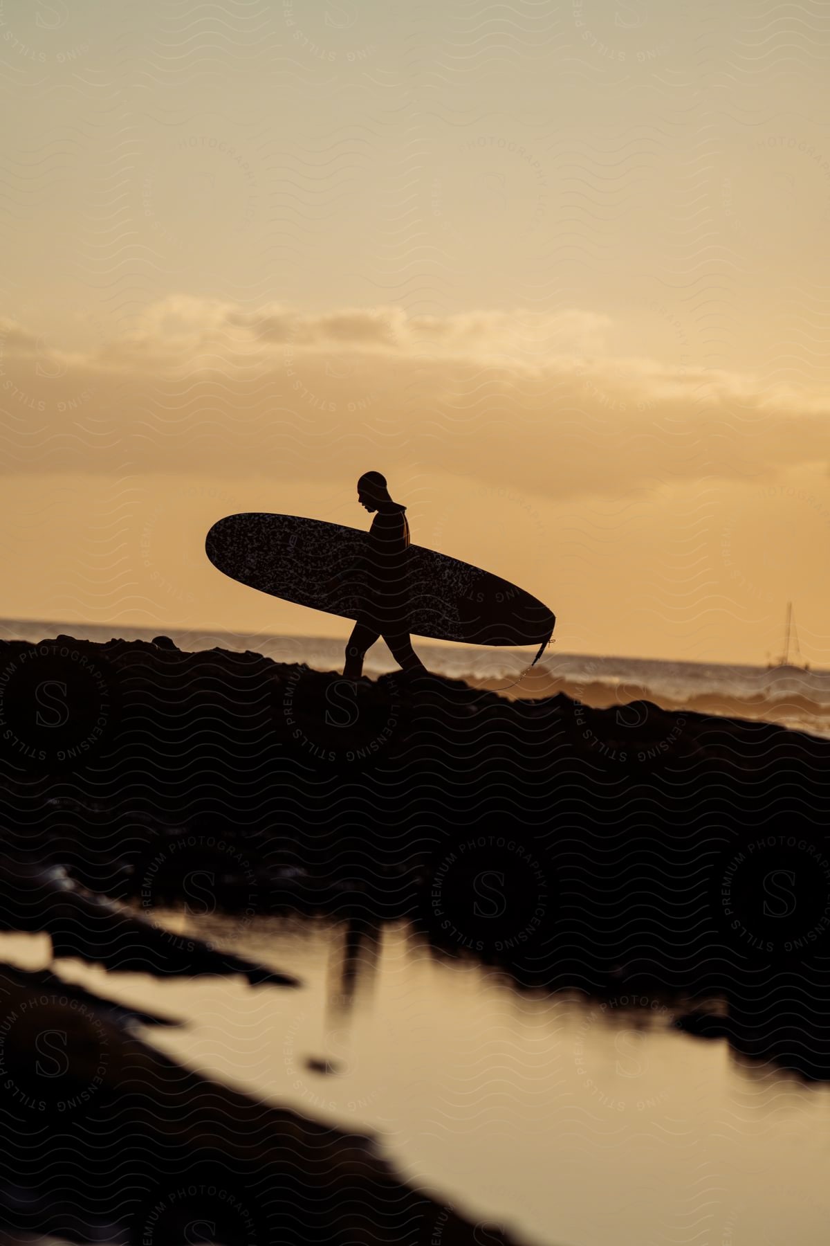 A person walking on a beach at sunset with a surfboard and a sailboat in the horizon