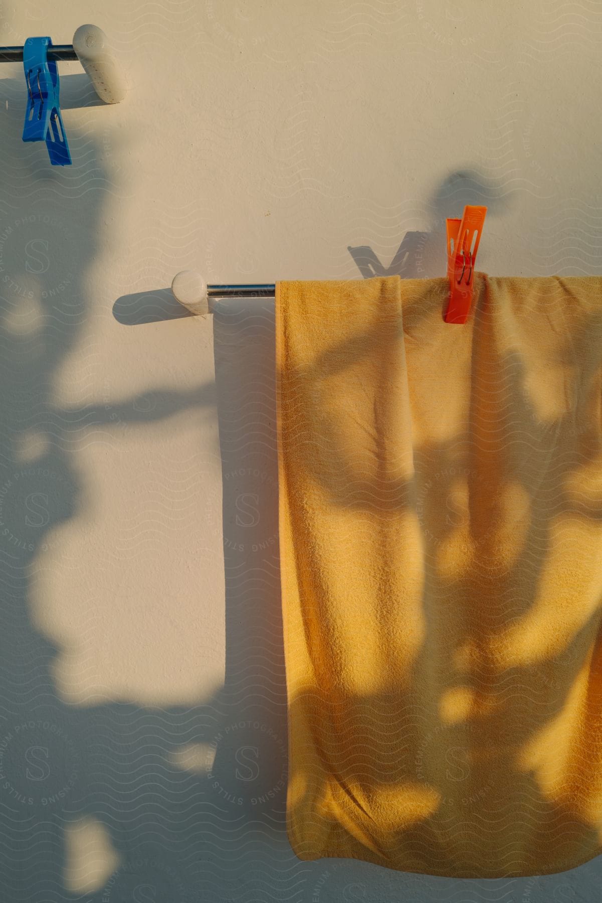 An orange towel hangs sideways on a clothesline dappled with shadows from tree branches against a wall