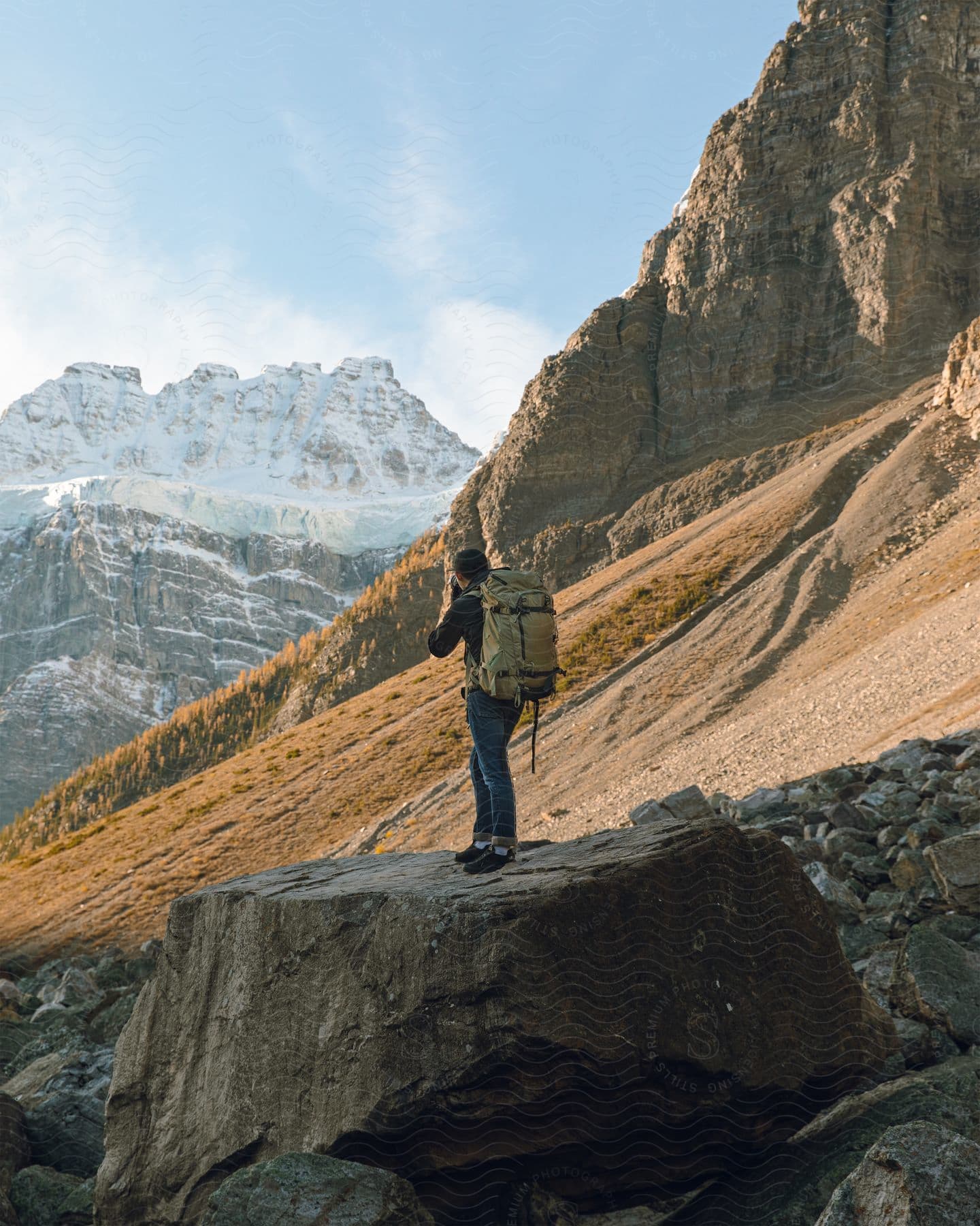 A hiker wearing a black jacket jeans and a brown backpack photographs snowy rugged mountains while standing on a rock