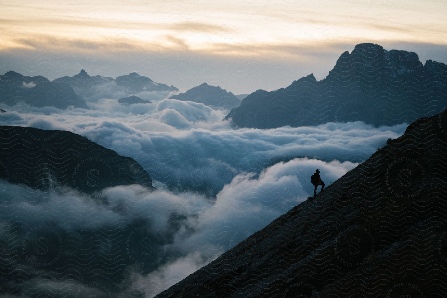 A person stands on a mountain slope next to lowhanging clouds at sunrise