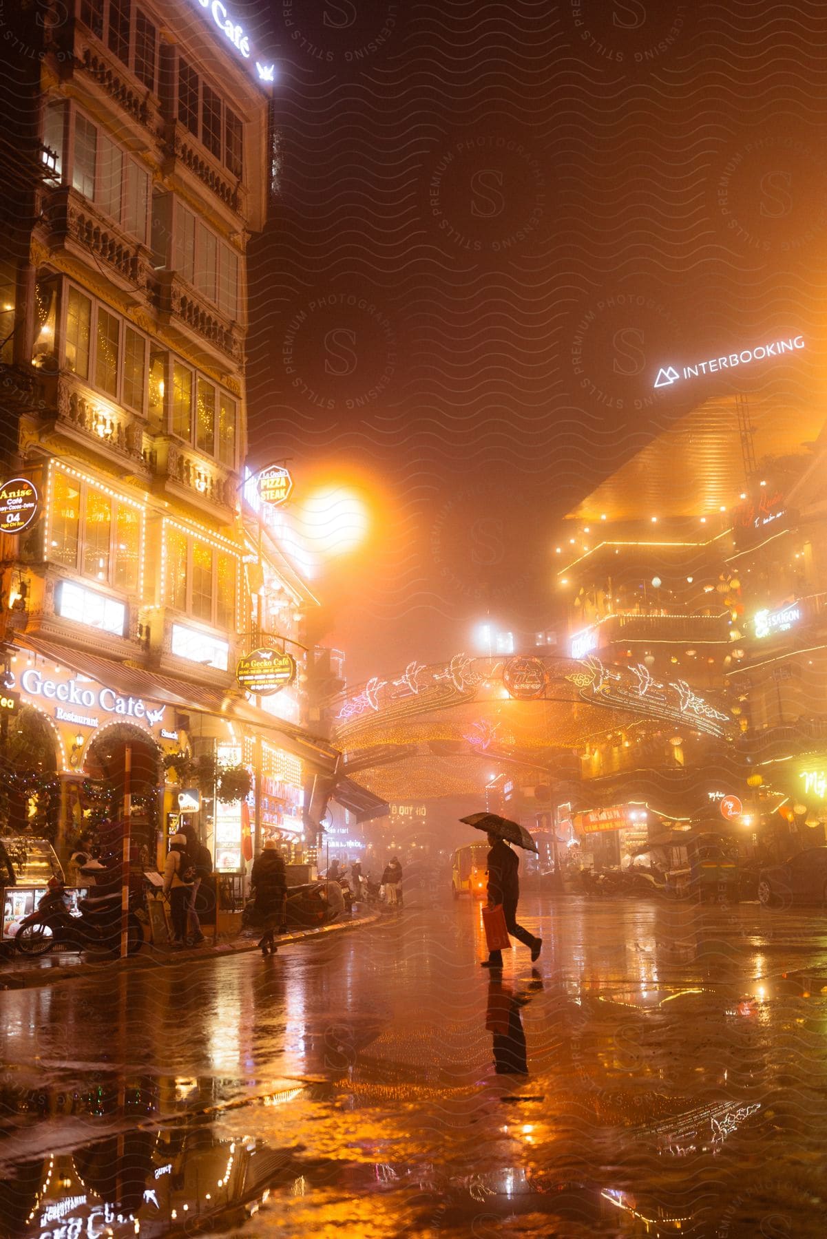 A person walks with an umbrella as gold lights reflect off a wet road from illuminated buildings on a rainy night in the city