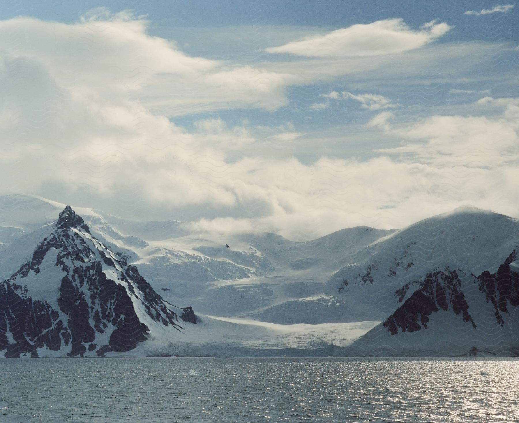 A snowcovered mountain range alongside a large body of water under mostly cloudy skies