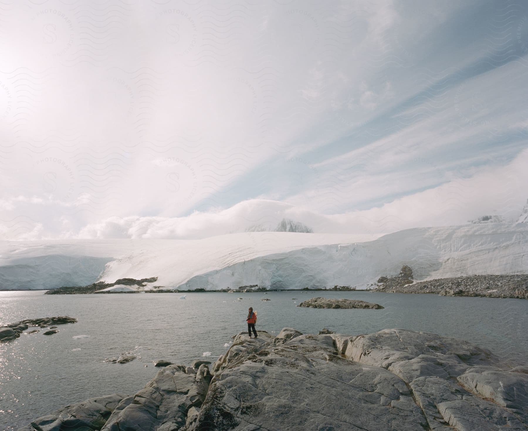 A hiker stands on a rocky shore across water from snow dunes