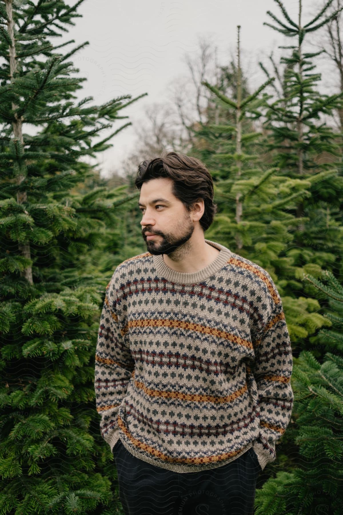 Man with beard in black pants and hands in pockets stands among trees