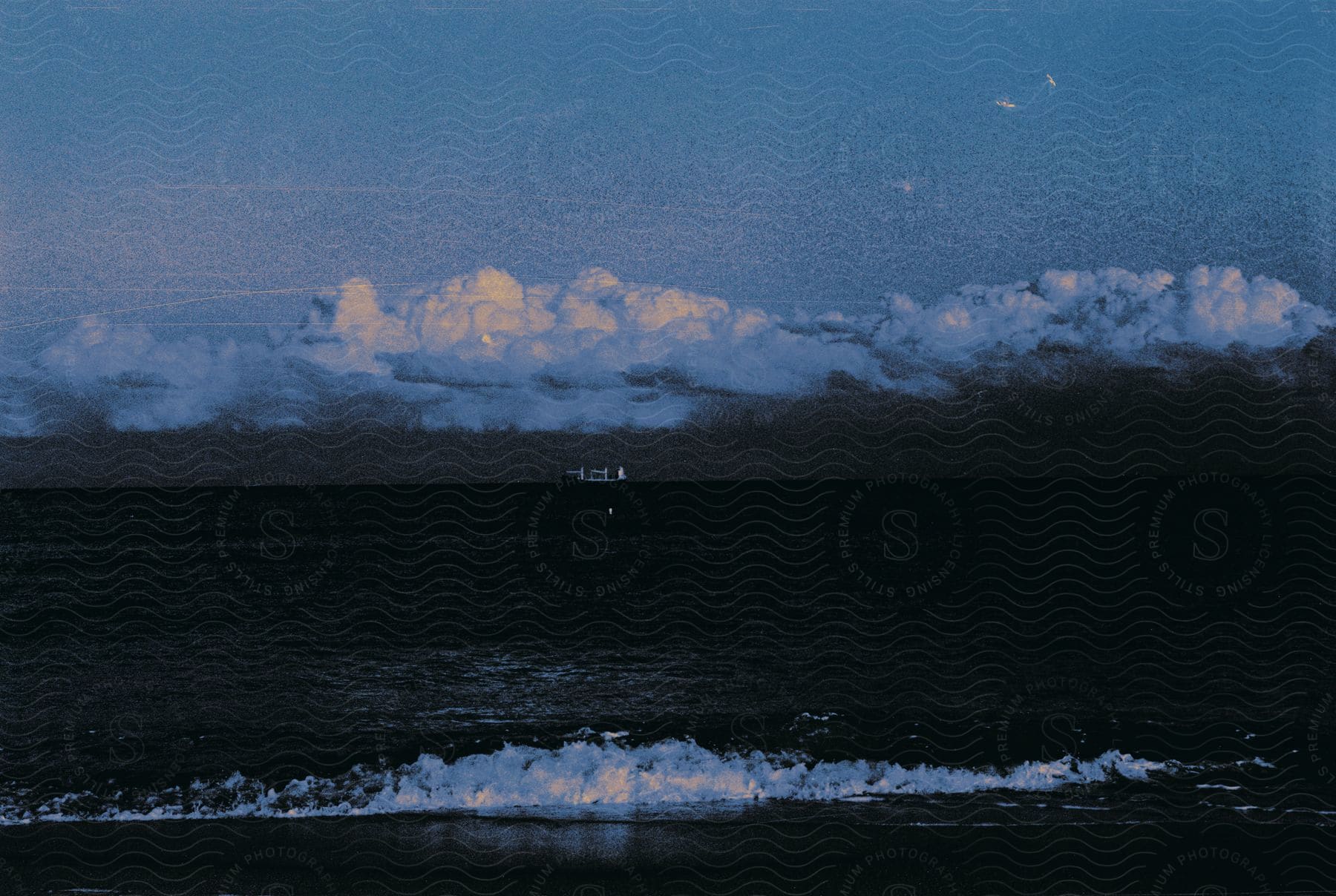 A boat is seen far away against a blue cloudy sky at dusk on a wide open ocean