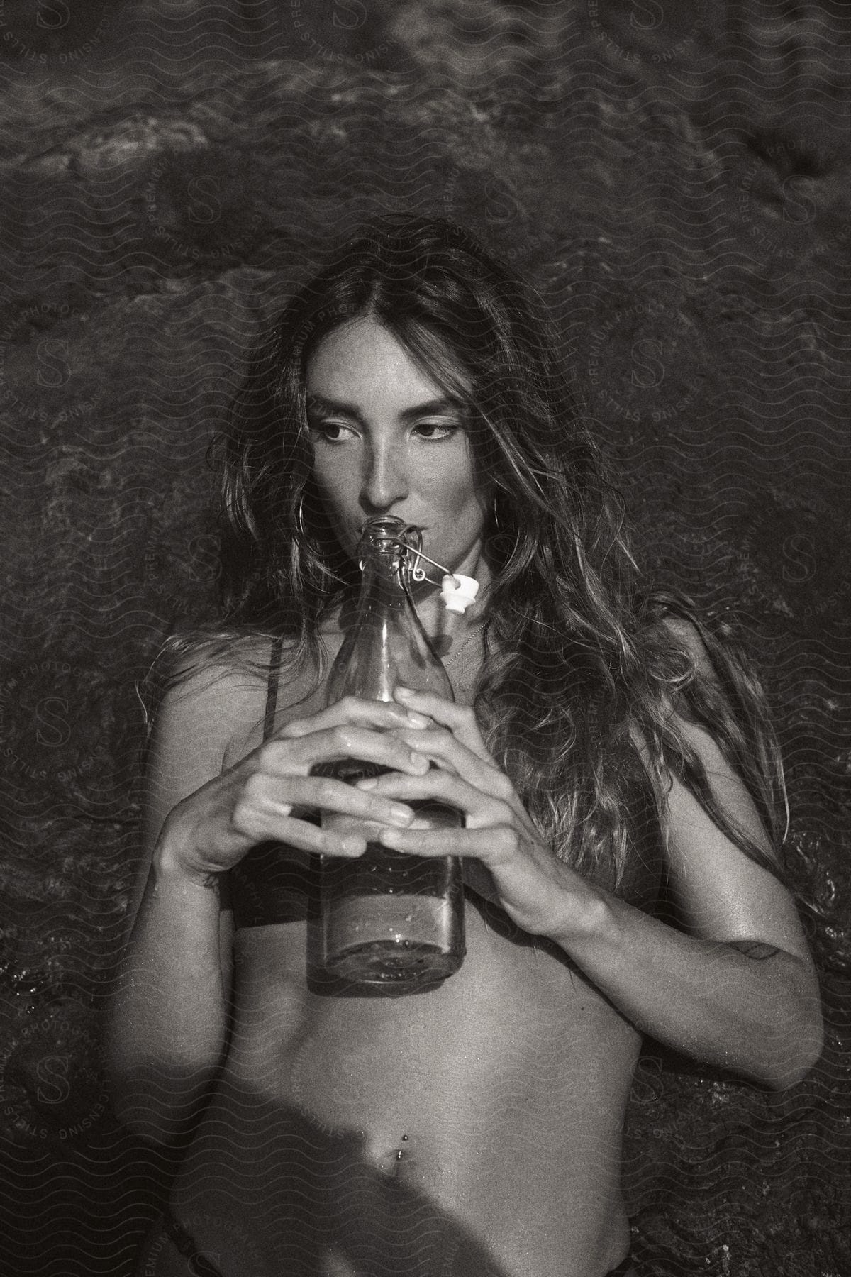Woman with brown wavy hair and bra lying down on rock holding a glass bottle