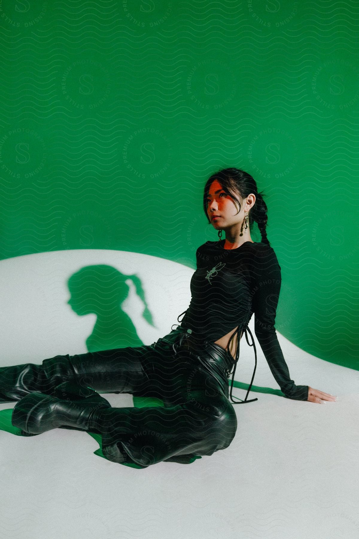 A young woman in black leather pants and platform shoes sits on a white surface in a green room