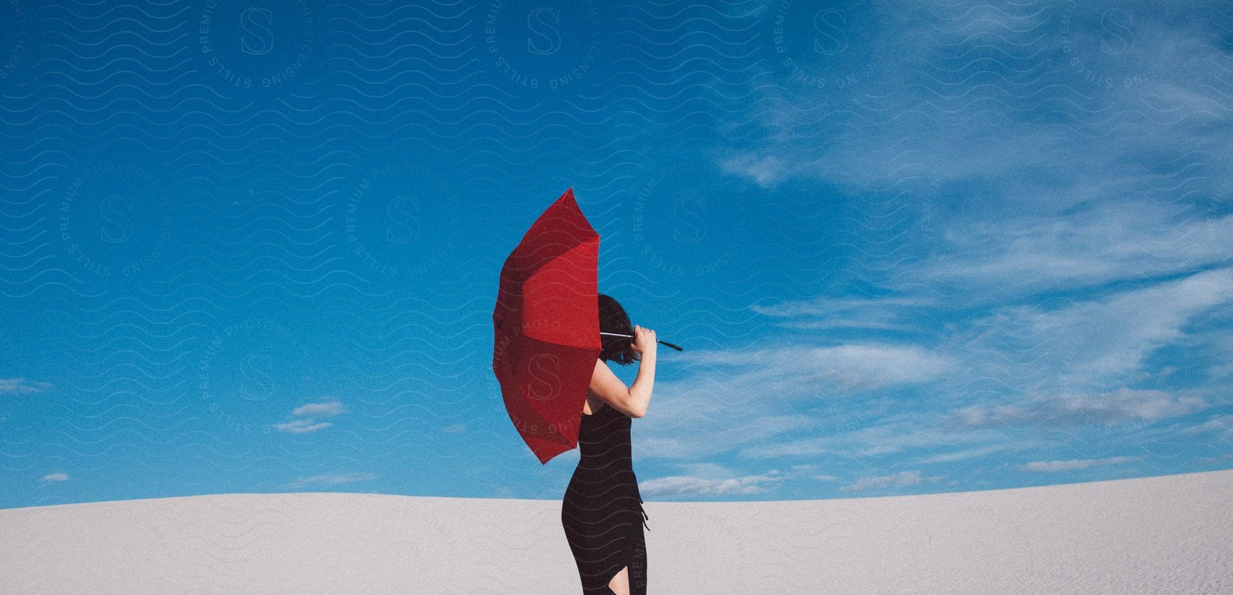 A woman in a black dress holds a red umbrella behind her head looking out over a vast white sand hill reaching towards a bright blue sky