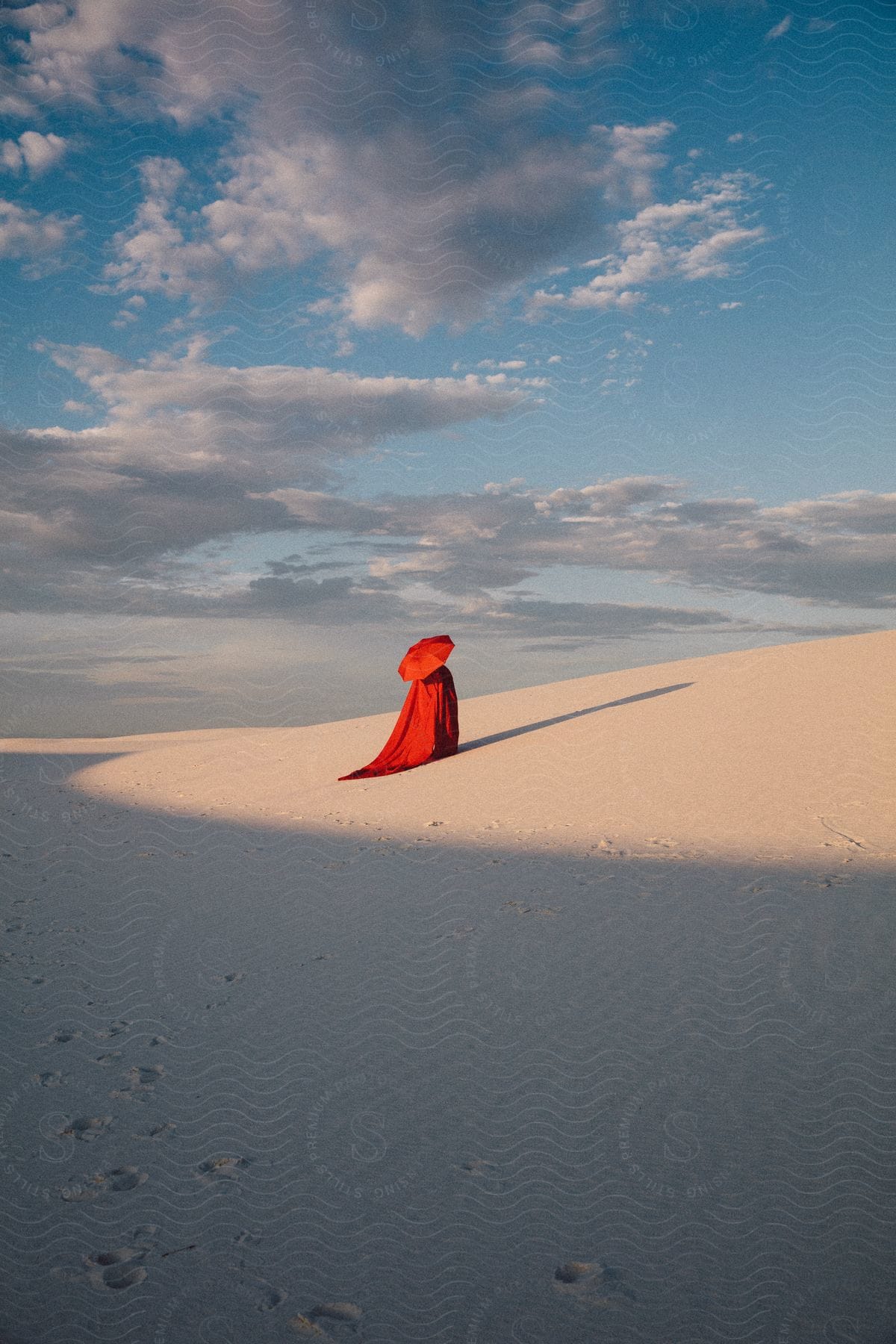 A person stands on a white sand dune hill under a blue sky holding a red umbrella