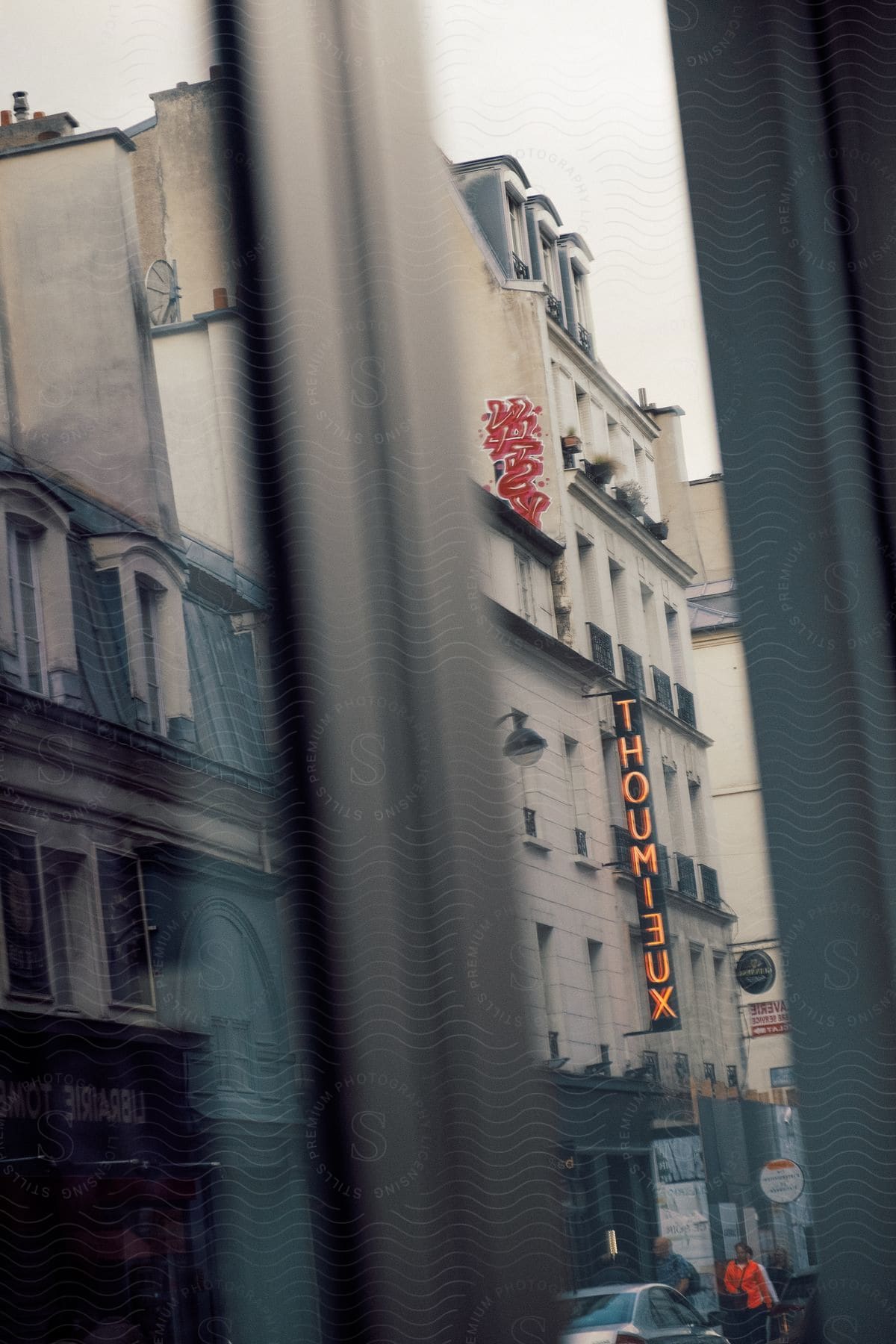 A hotel window overlooks a street filled with parked cars neon signs and people walking by