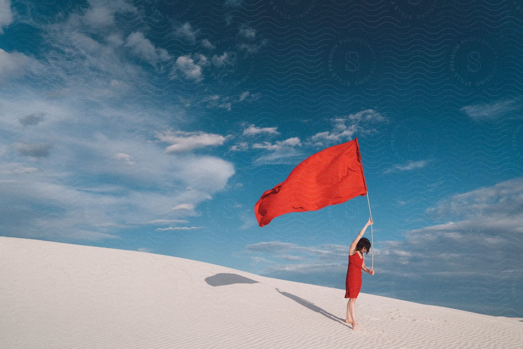 A person stands alone under a red flag on a desert slope with a clear blue sky