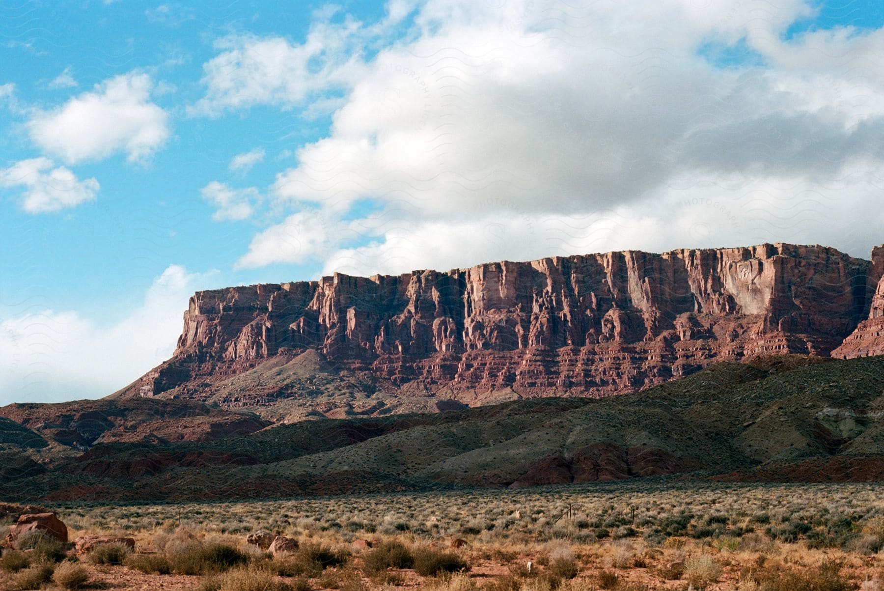 Rolling hills in front of red rock plateaus in rainbow canyon arizona under white clouds in a blue sky