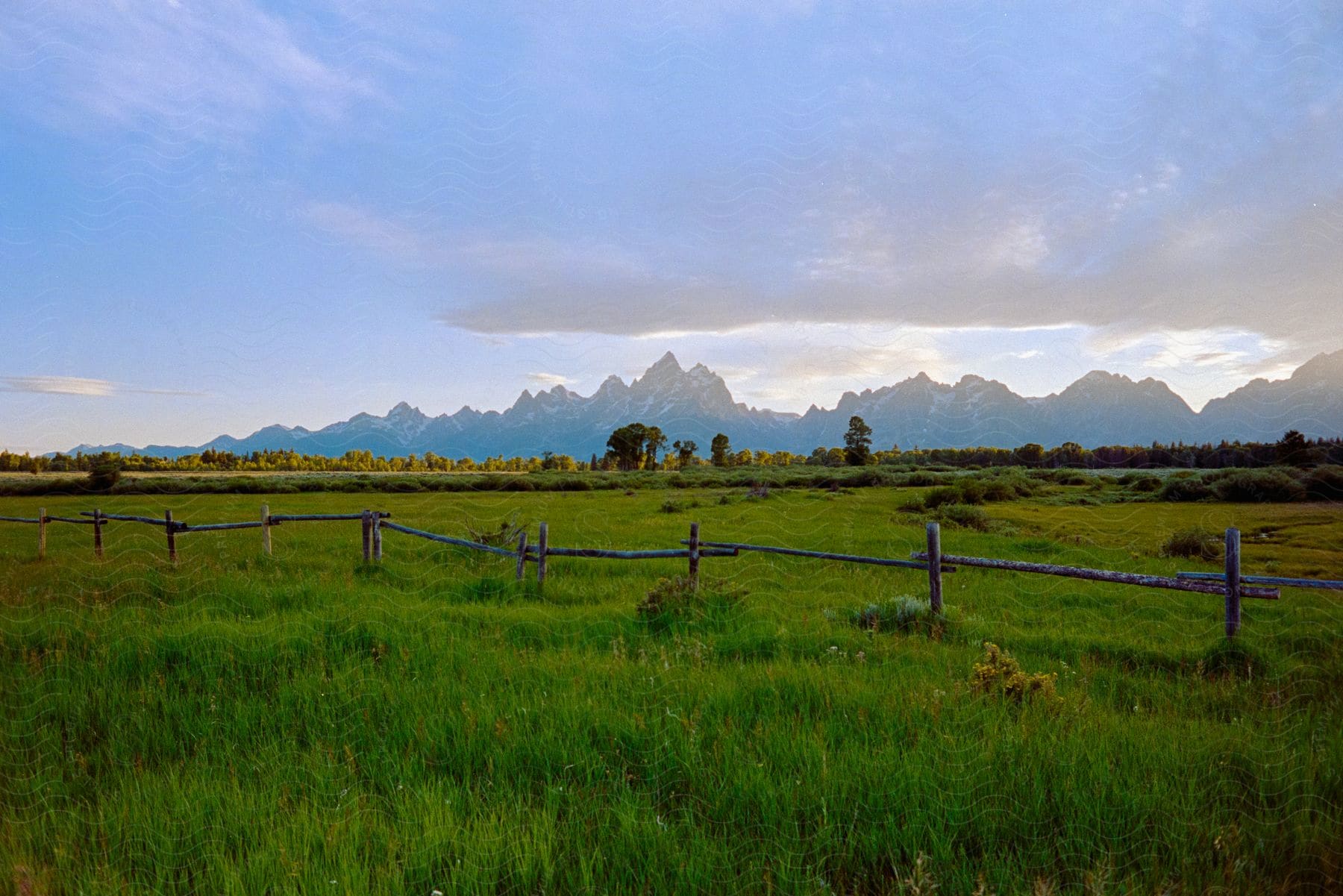 A post and rail fence runs along a grassy field with trees and mountains in the distance