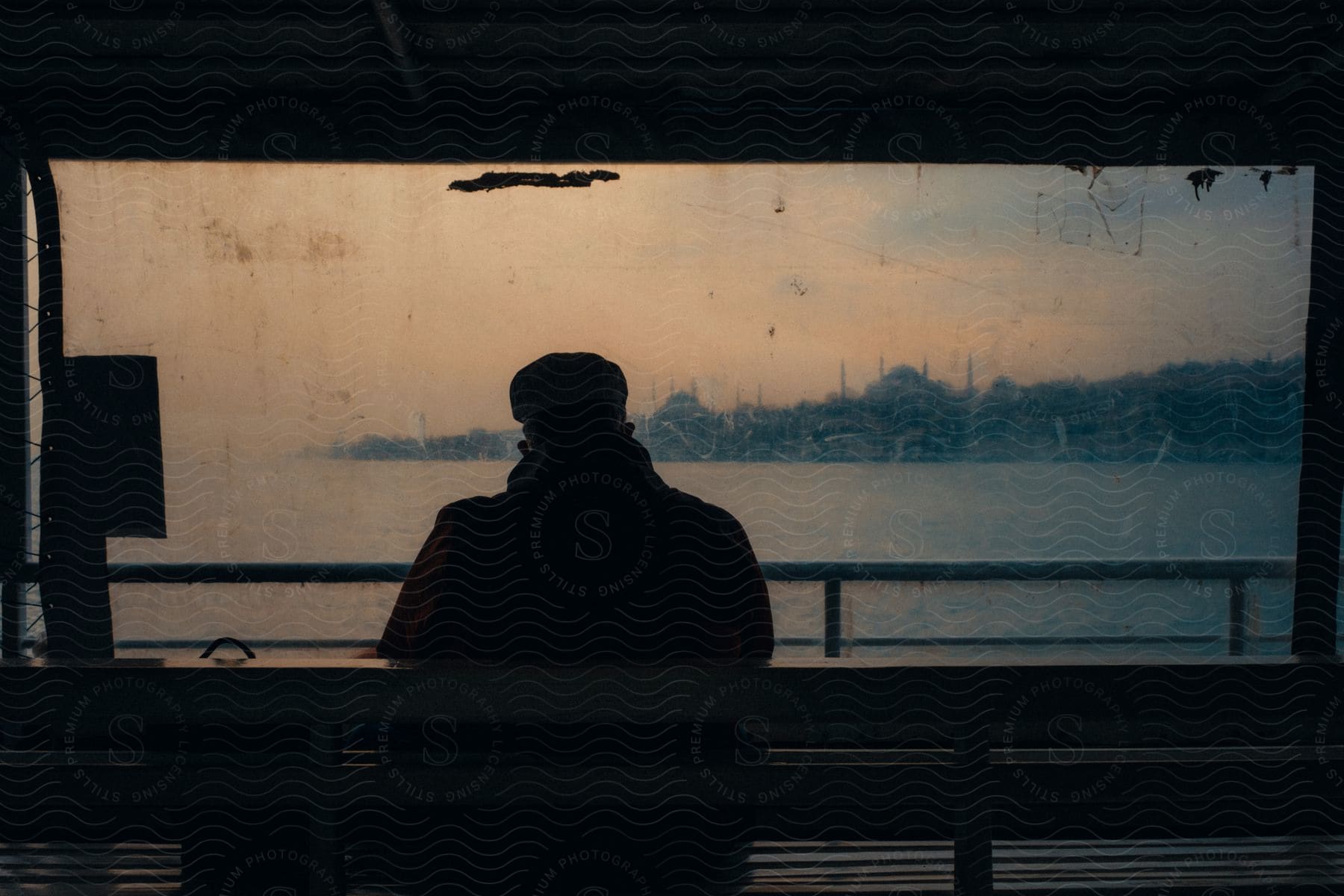 A man in a coat and cap sits on a ferrys bench looking at a dirty window