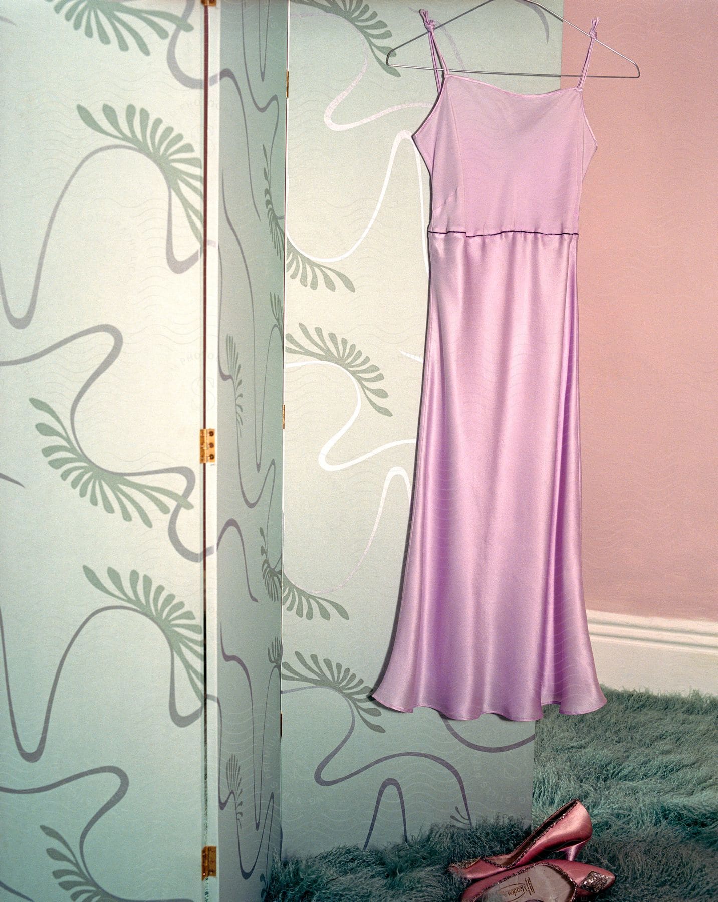 Pink dress hanging on a hanger with pink high heels placed underneath on the floor in a dressing room