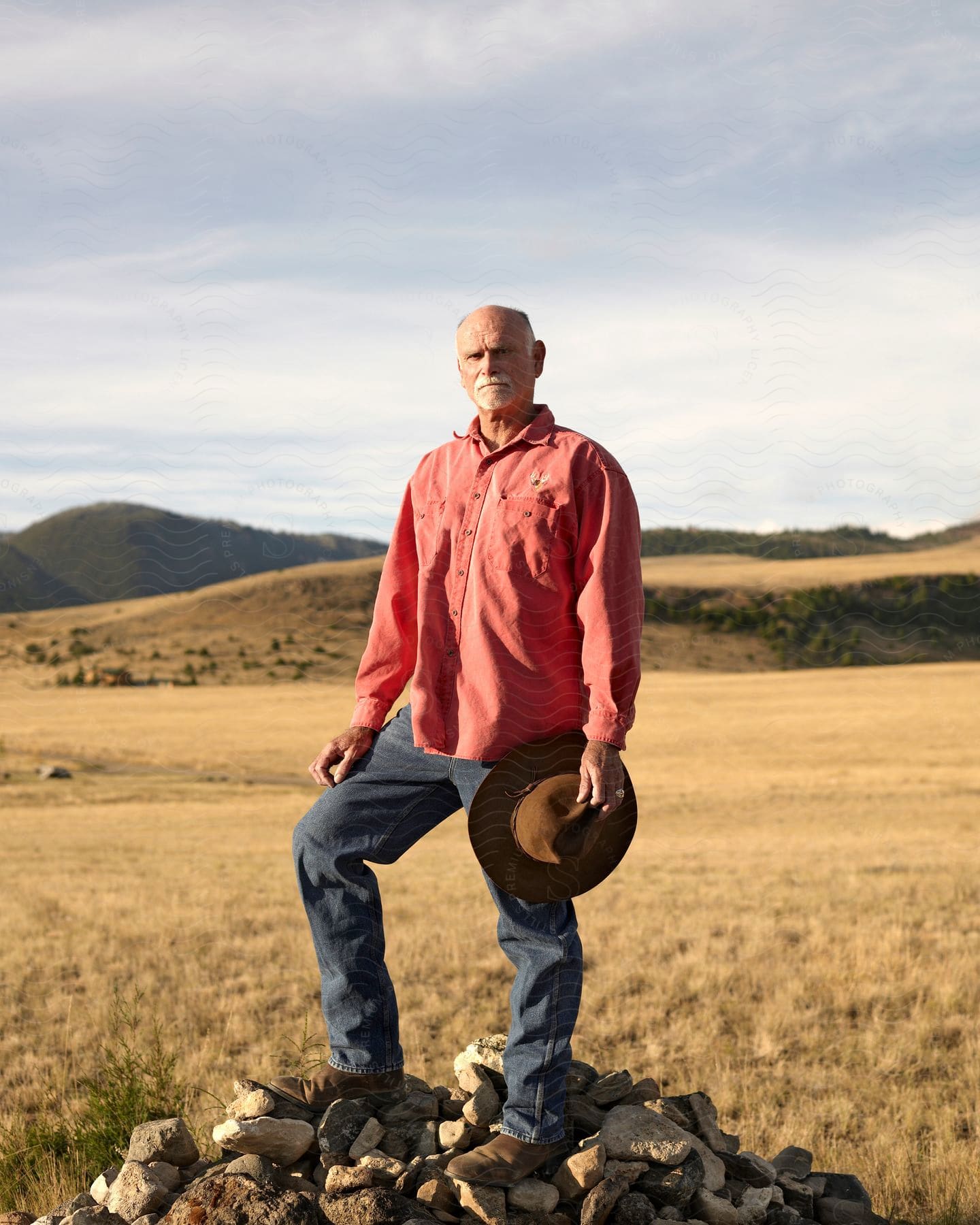 Lone cowboy atop rock pile in dry grass field holding hat surveying landscape