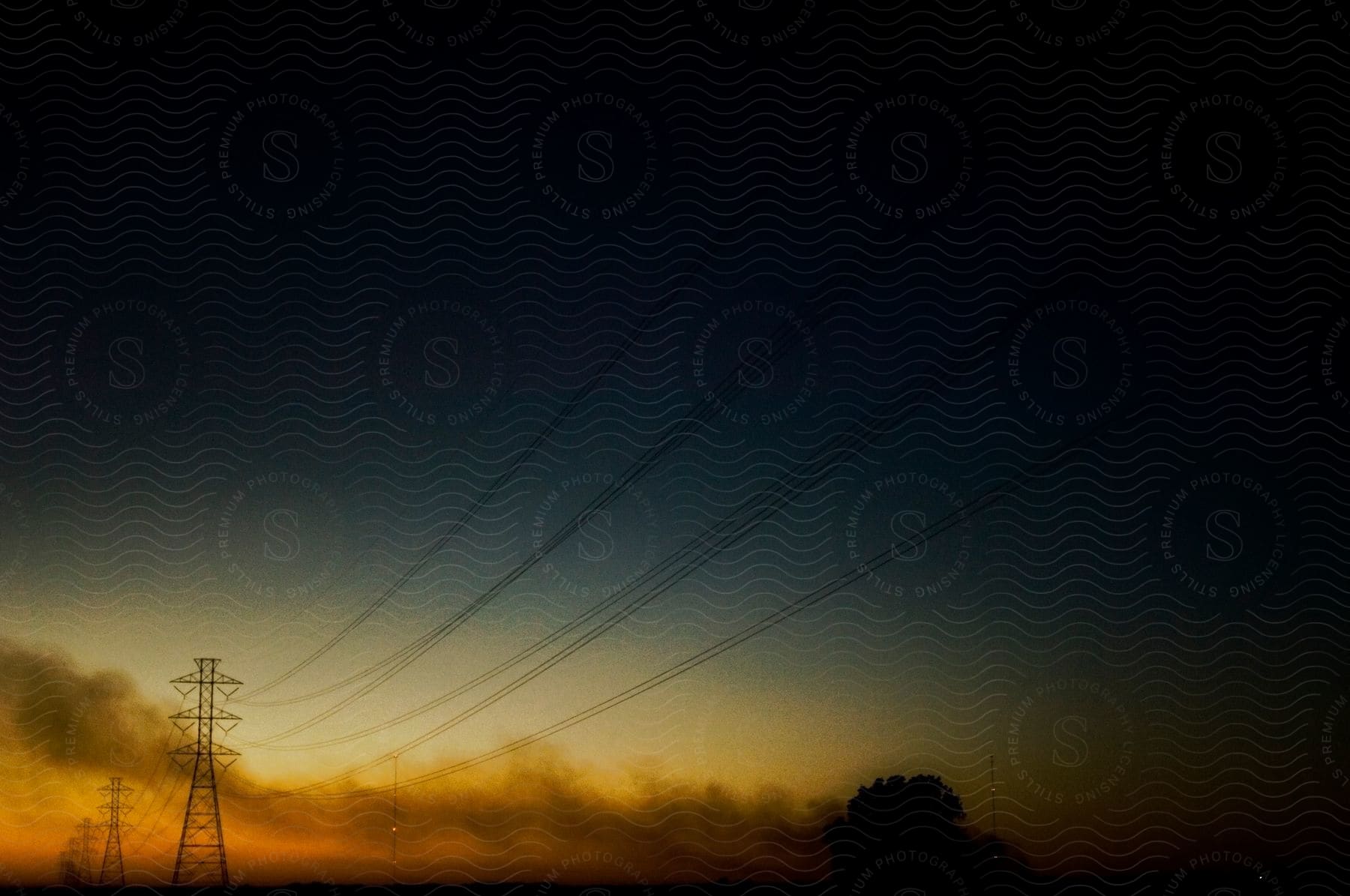 A lone tree stands amidst power lines and smoke as the sun sets in the background