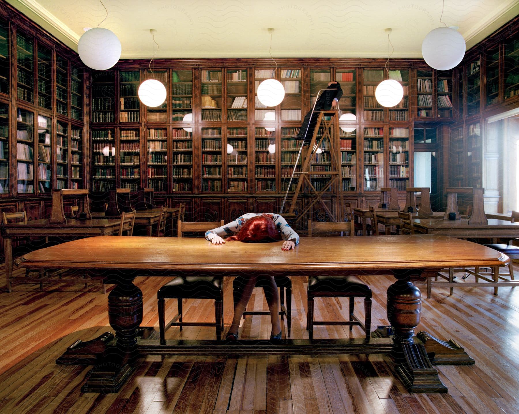 A man sleeps on a ladder behind a woman asleep at a table in a library