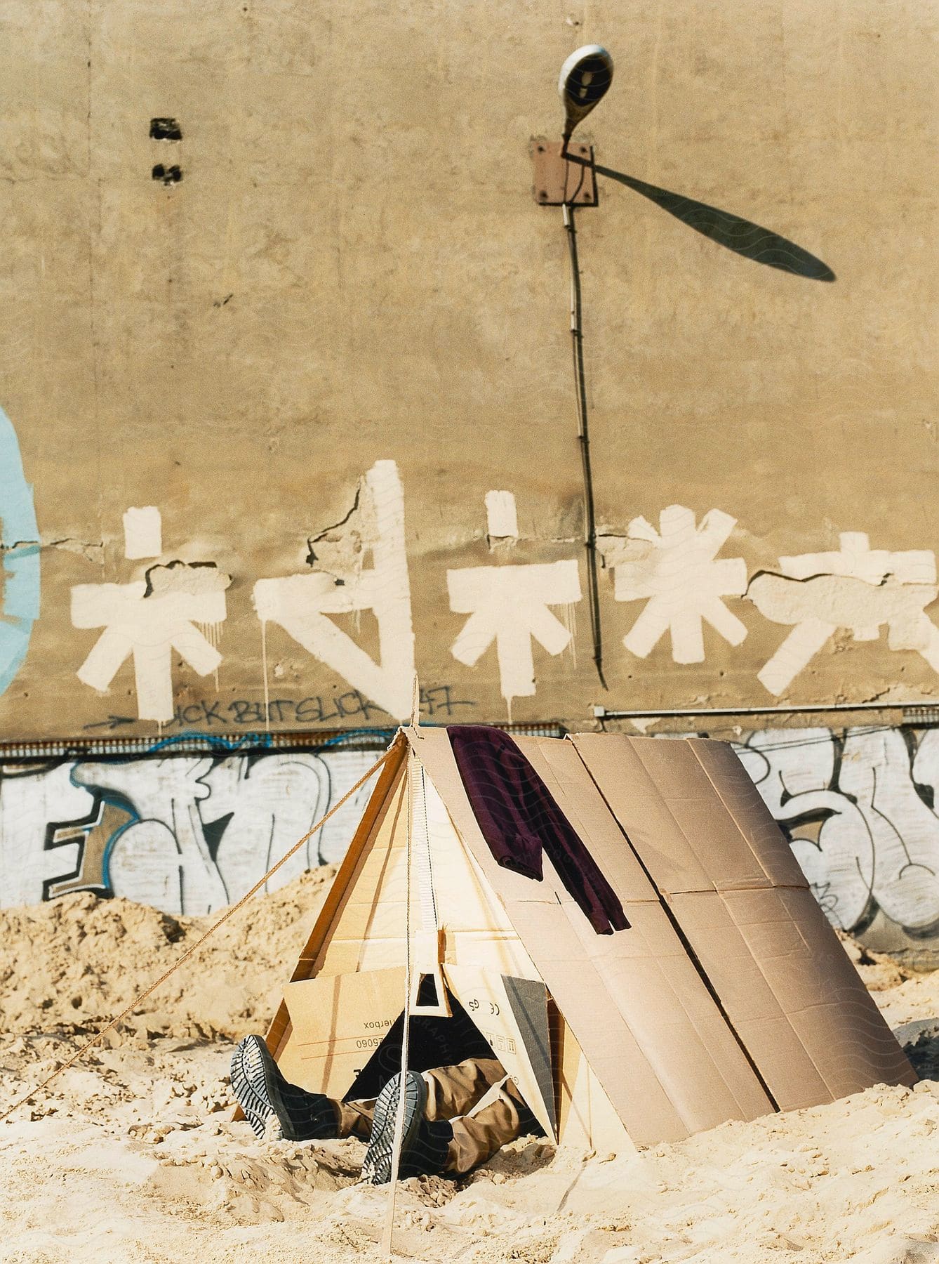 Person wearing black boots inside a cardboard tent next to a graffiticovered wall and street lamp