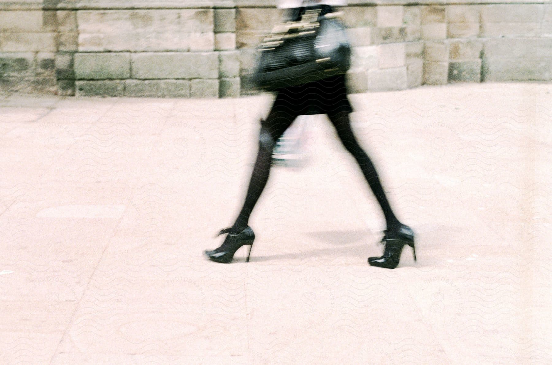 A woman in black clothing and high heels walks on a sidewalk in front of a stone building carrying a black purse