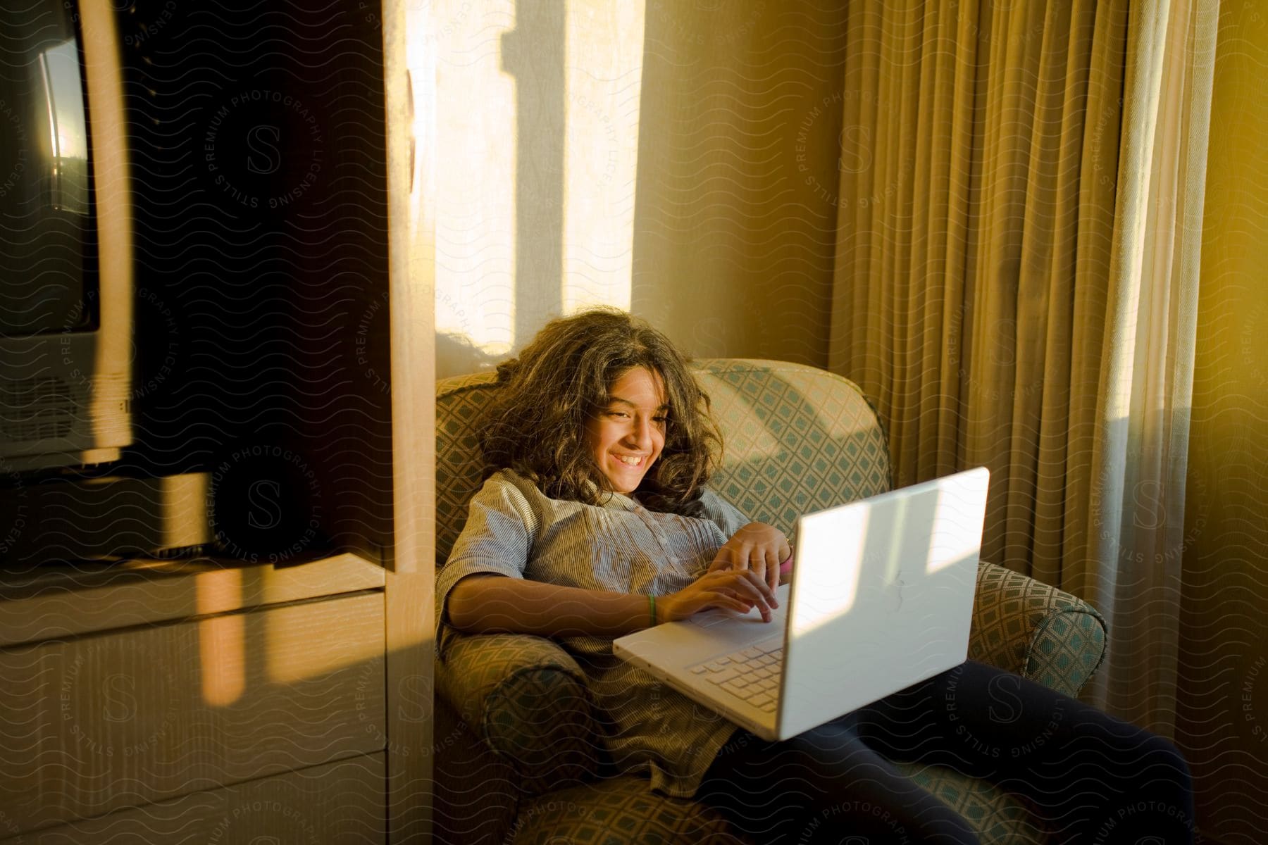 A girl sits on an armchair next to a tv and smiles while using a laptop in a room with sunlight coming through the curtain