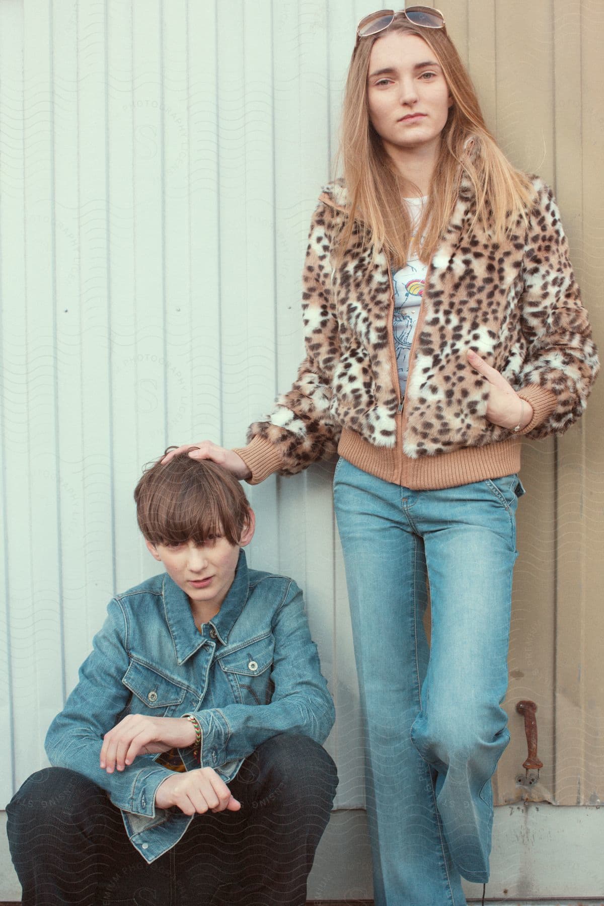Stock photo of teenage girl modeling animal print jacket and blue jeans standing against panel siding building with male teenage model beside in blue jean jacket