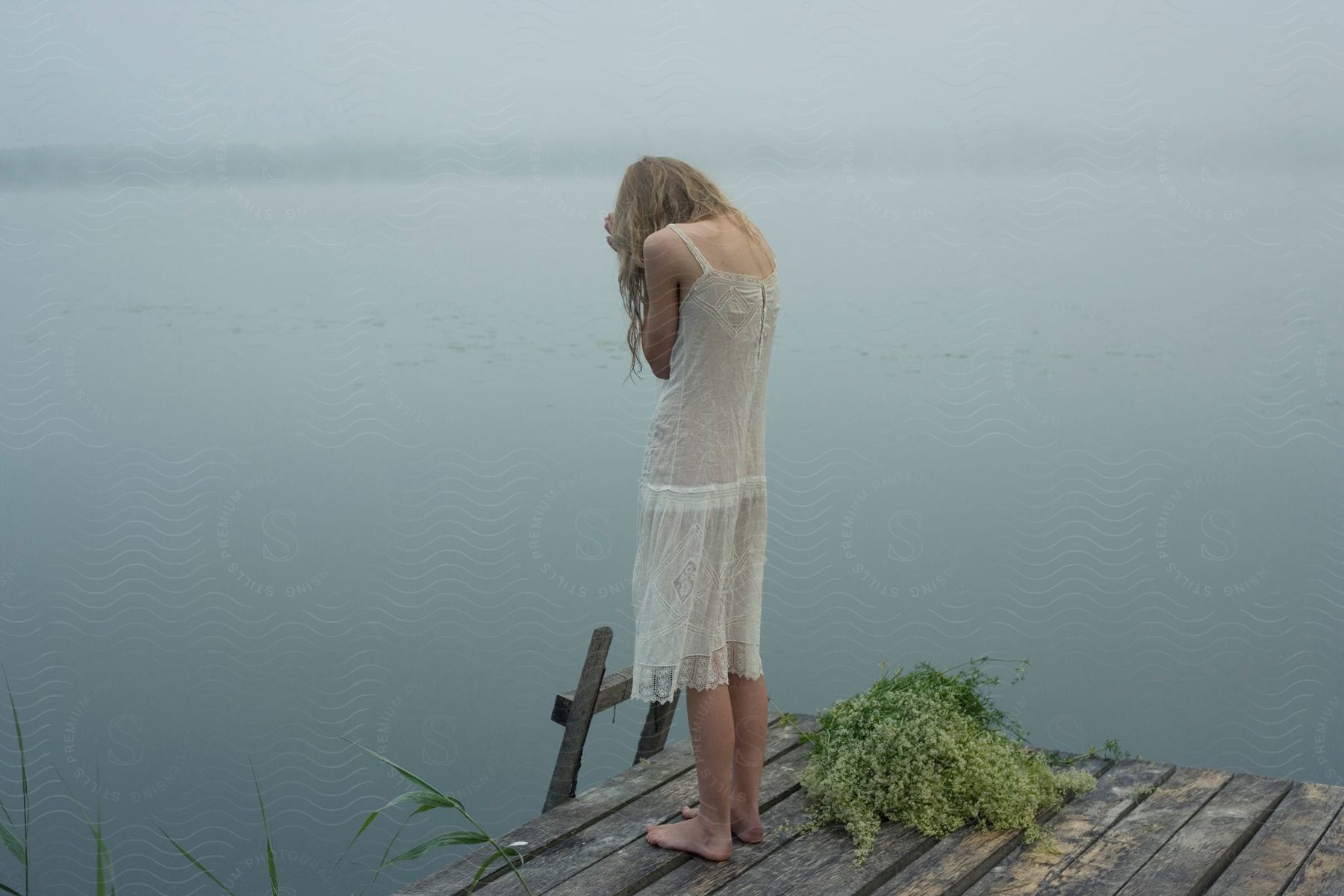 A blonde girl standing on the end of a wooden pier wearing a sheer shift dress and looking down