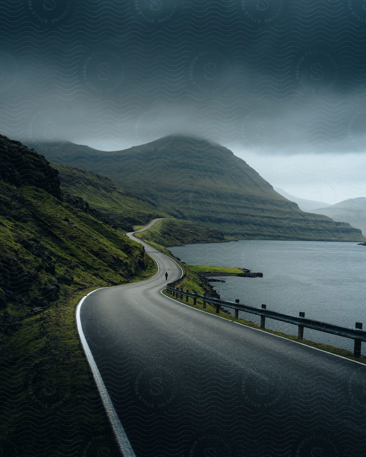 Person walking on a rainy road with water on one side and mountains obscured by clouds on the other