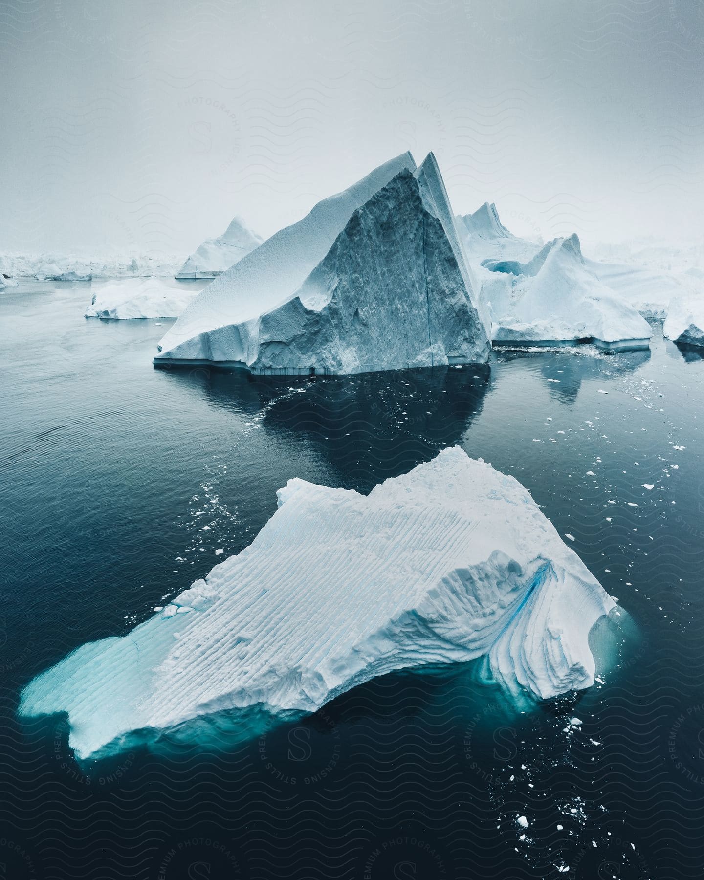 Icebergs and pieces of ice float in arctic water under a hazy white sky